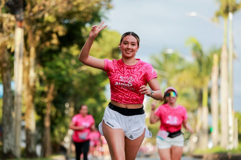 Energetic woman running outdoors in pink event shirt, smiling and flashing a peace sign.