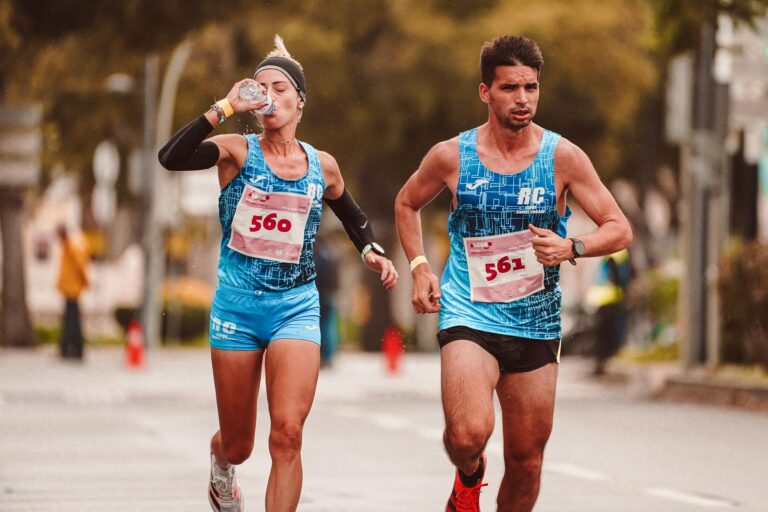 Two athletes participating in a marathon, hydrating and focused, while running outdoors on a city street.